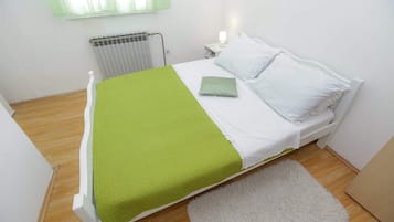 Una cama con colchón blanco y ropa de cama verde, una mesita de noche blanca con lámpara, un radiador y una alfombra blanca sobre piso de madera.