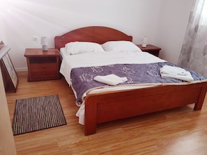 A neatly made bed with a purple blanket and white pillows, a wooden headboard, a bedside table with a lamp, and a small rug on a wooden floor. - Apartment 'Family Apartments' with Sea View, Wi-Fi and Air Conditioning (Pakoštane)
