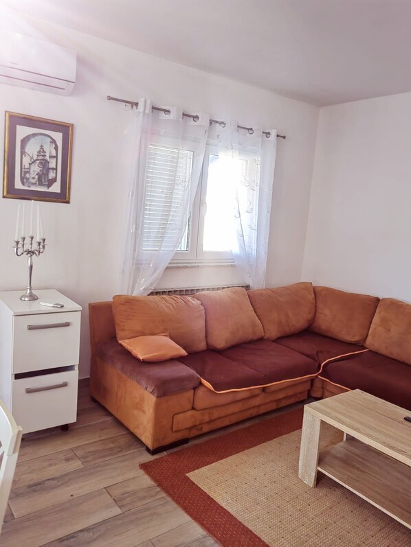 A living room with a brown sofa, a white side table, a small wooden table, a candelabra, and a window with sheer curtains. - Apartment 'Family Apartments' with Sea View, Wi-Fi and Air Conditioning (Pakoštane)