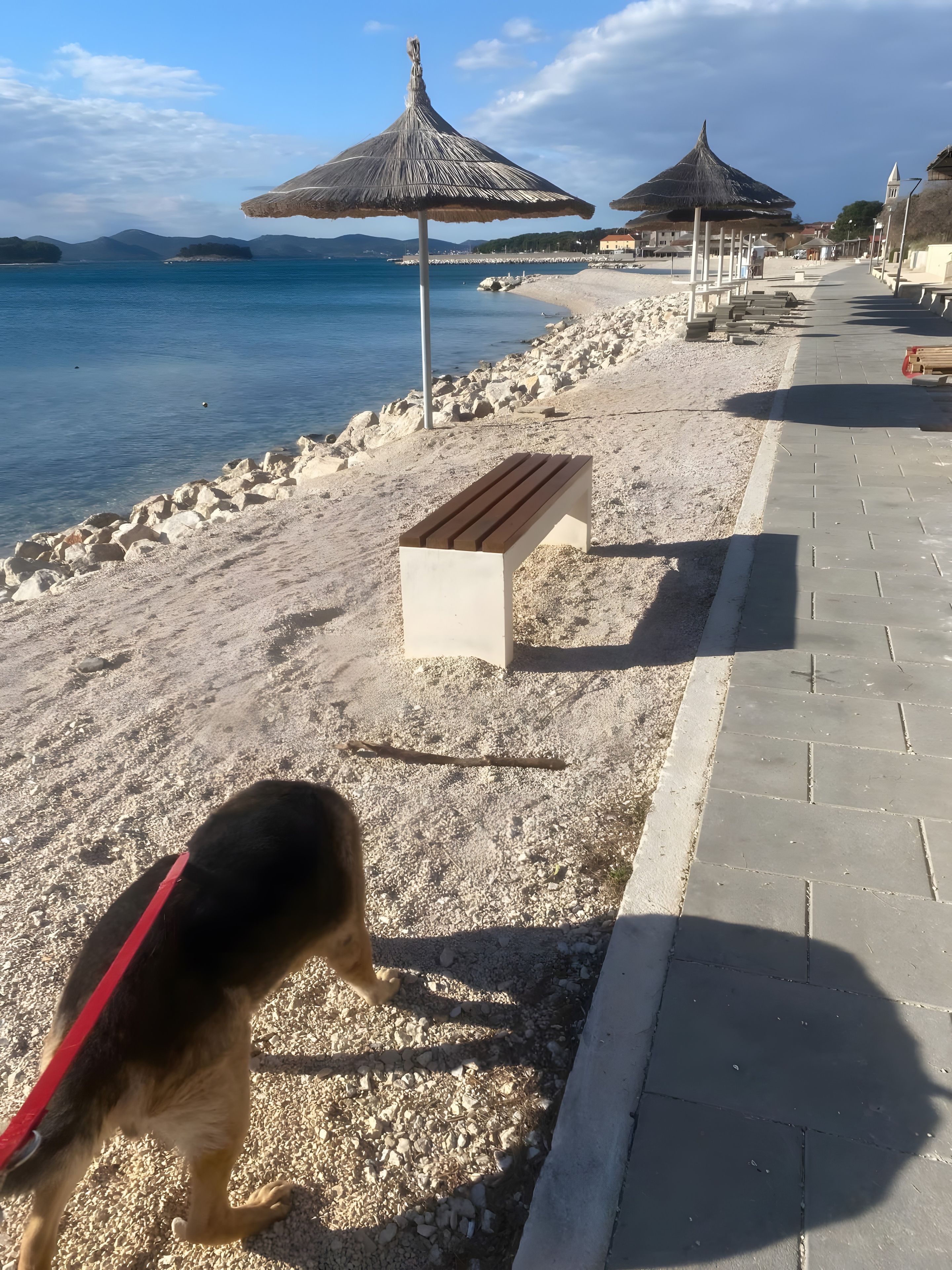 A beach with a pebble path, a wooden bench, and a dog on a leash.