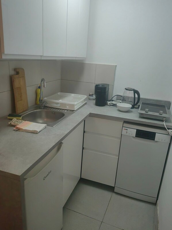 A compact kitchen with a dishwasher, a kettle, a coffee maker, and a cutting board.