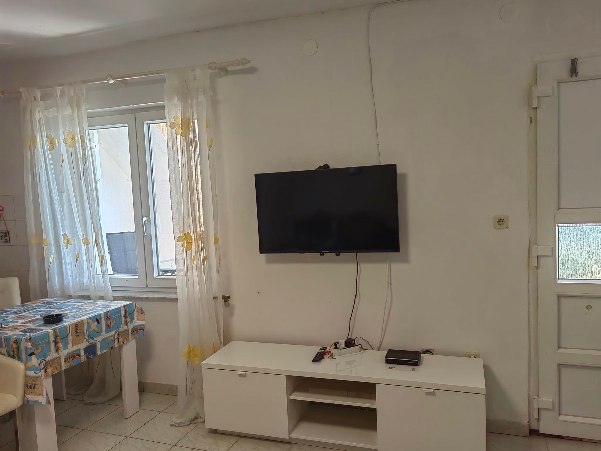 A room with a television mounted on the wall, a white desk with a computer, a table with a patterned tablecloth, and a window with curtains.