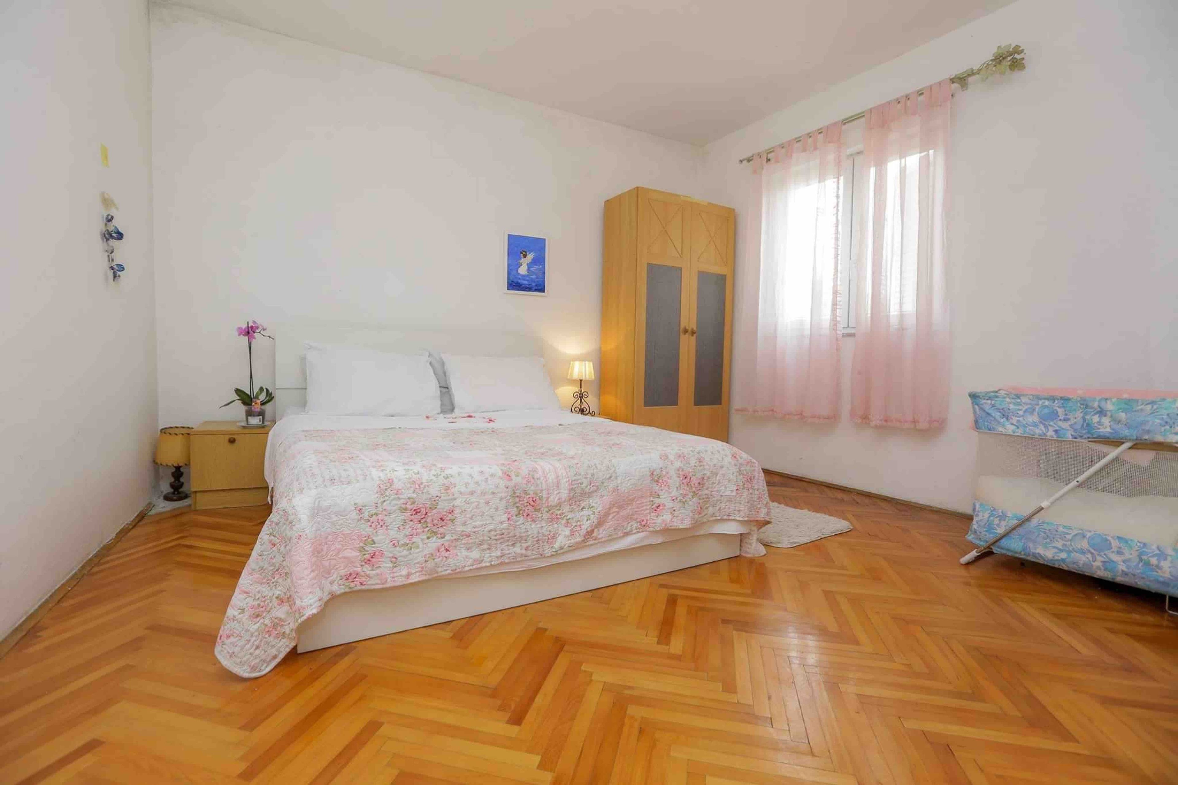 Ein Schlafzimmer mit Holzboden, einem Bett mit floralem Überwurf, einem Kleiderschrank, einem Nachttisch mit Lampe und einem Fenster mit durchsichtigen Vorhängen.