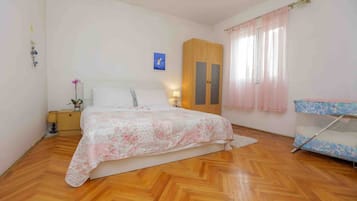 Ein Schlafzimmer mit Holzboden, einem Bett mit floralem Überwurf, einem Kleiderschrank, einem Nachttisch mit Lampe und einem Fenster mit durchsichtigen Vorhängen.