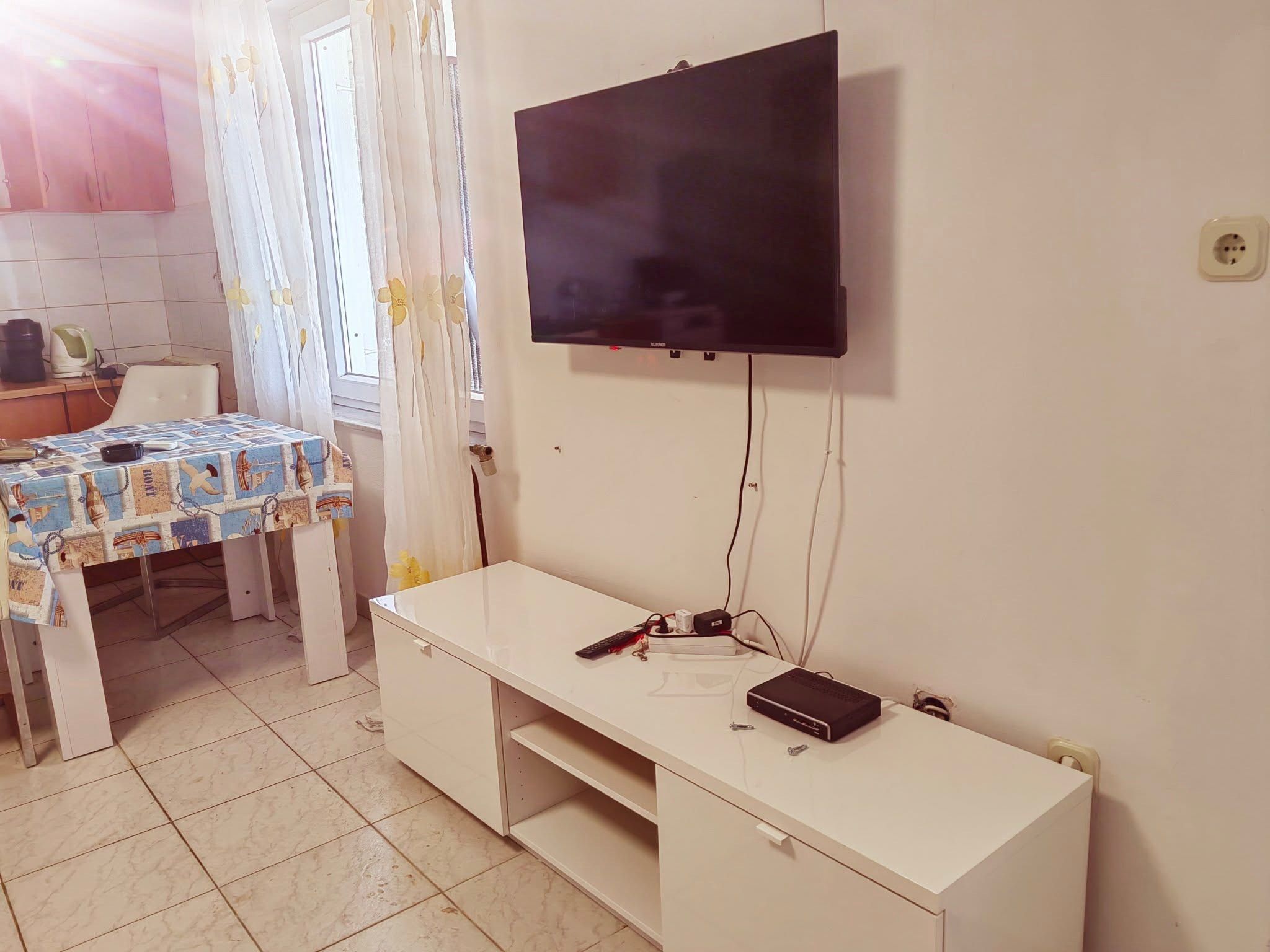 Une pièce avec un téléviseur fixé au mur, un bureau blanc avec un ordinateur et une télécommande, et une table recouverte d  ’une nappe à motifs.