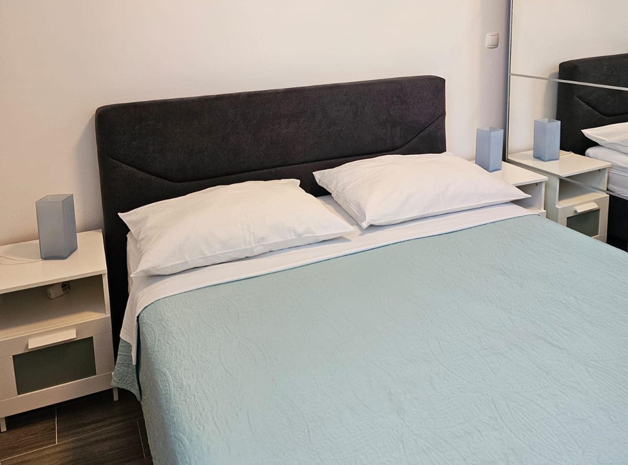 Ein ordentlich gemachtes Bett mit weißer Bettwäsche und einer hellblauen Decke, an beiden Seiten flankiert von weißen Nachttischen mit je einem Lautsprecher.