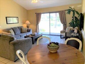 A living room with a sofa, a coffee table, a dining table with a fruit bowl, a television, and a plant. - Lake Views and Plenty of Room for All! (Palm Desert)