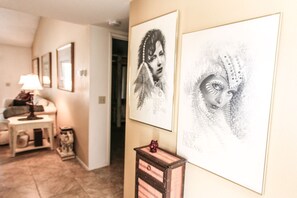 A hallway with framed artwork on the walls, a lamp on a side table, and a small cabinet with a decorative item on top. - Enjoy the Outdoors on the 16th Fairway! (Palm Desert)