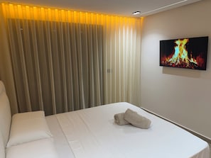 Quarto de hotel com cama, televisão exibindo uma lareira e janela com persianas verticais.