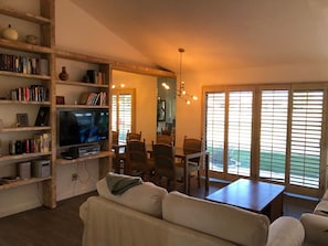 A living room with a television, bookshelves, a sofa, a dining table with chairs, and large windows with blinds. - Great location on 11th Fairway! (Palm Desert)