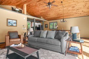 A cozy living room with a grey sofa, a brown leather chair, a wooden ceiling, and a dining area with a lamp and books. - Modern Alpine Chalet 2 Miles from Belleayre (Pine Hill)