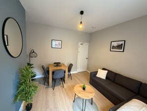 Una sala moderna con un sofá, una mesa de comedor con sillas, una pequeña mesa redonda con una planta, una maceta y dos cuadros en la pared.