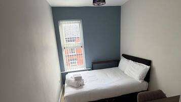 A small, modern bedroom with a single bed, a small table, and a window with a view of a building.