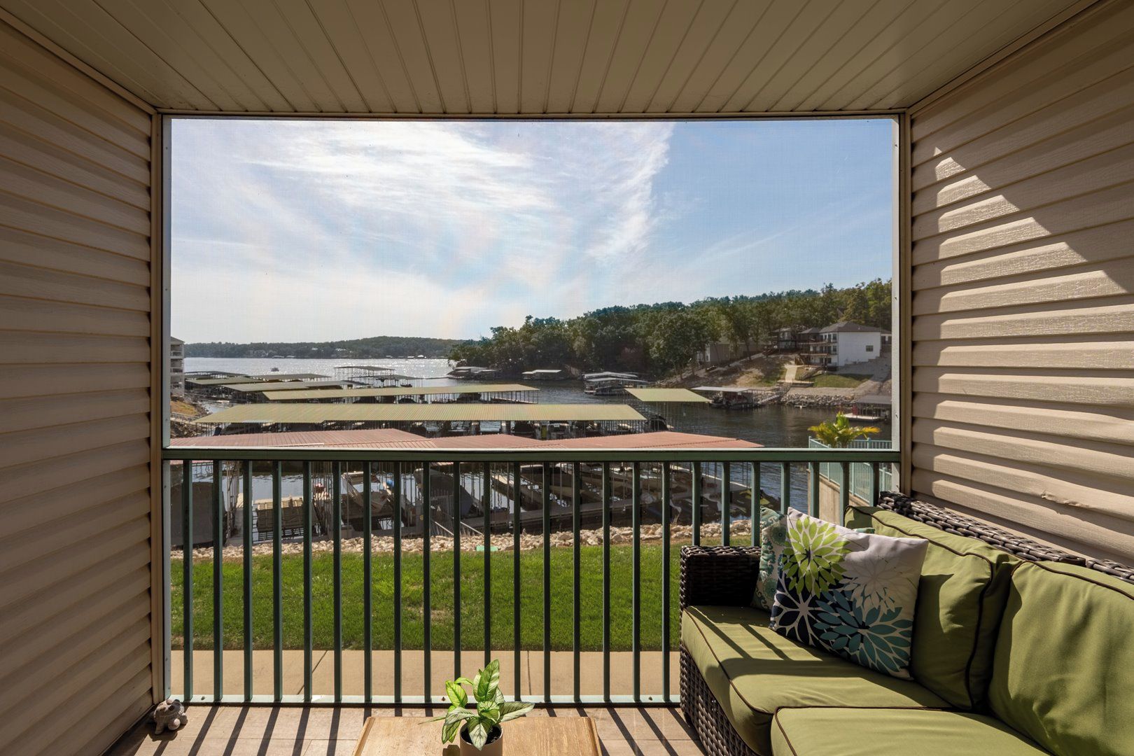 A balcony with a view of a waterfront, a green lawn, and a seating area with cushions.