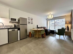A compact kitchen with a microwave, refrigerator, and dishwasher. There is a dining area with a round table and chairs. A sofa and a lamp are visible in the living area. - 300m from ski slopes, 1st floor, terrace, tv, ski locker, 58m², Serre Chevalier (Briançon)