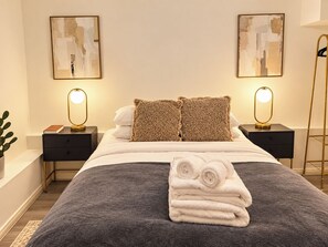 A neatly made bed with a grey blanket, two pillows, and a stack of white towels. There are two bedside tables with lamps, a floor lamp, and two abstract paintings on the wall. - Compact Split-Level Studio near Paddington (Greater London)