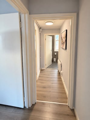 A hallway with wooden flooring, white walls, and a door leading to a bathroom. - Cosy One-Bedroom Flat near Paddington (Greater London)