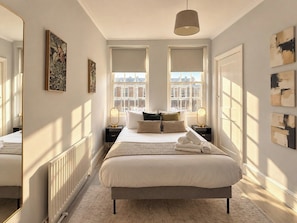 A bedroom with a large bed, two bedside tables, a radiator, and a view of the city through windows. - Cool Soho Room Steps from Chinatown & West End (Greater London)