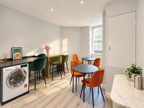A compact kitchen with a washing machine, a dining area with chairs, and a small table. - Soho Hideaway | Private Room near West End (Greater London)