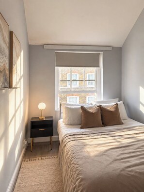 Un dormitorio con cama, mesita de noche, lámpara y ventana con vistas a edificios.