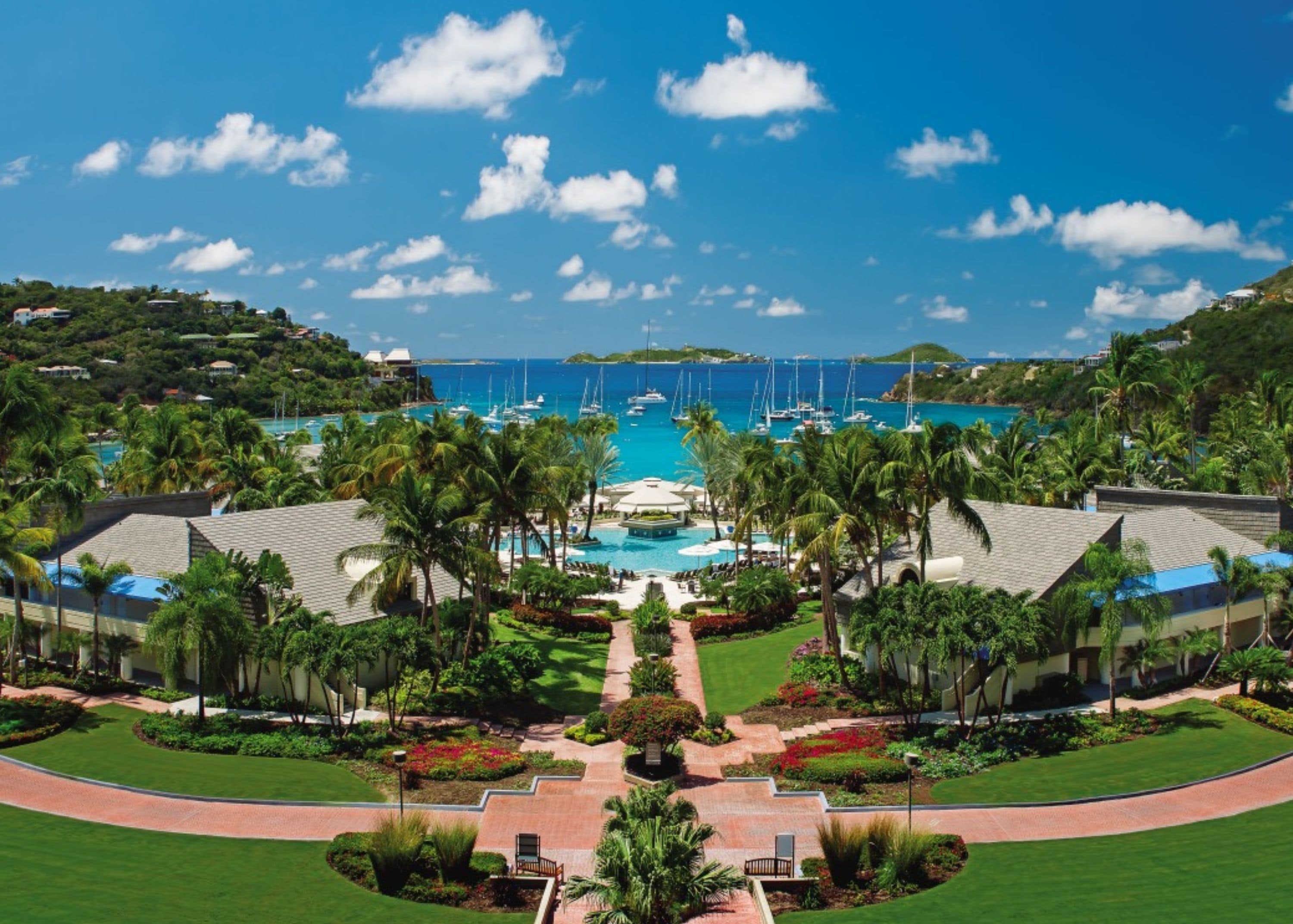 A resort with a central pool, surrounded by palm trees and a clear view of the ocean.