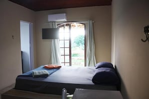 A single bed with a flat-screen TV mounted above it, a window with curtains, and a door to the left. - Sítio Bom Retiro (São Pedro da Aldeia)