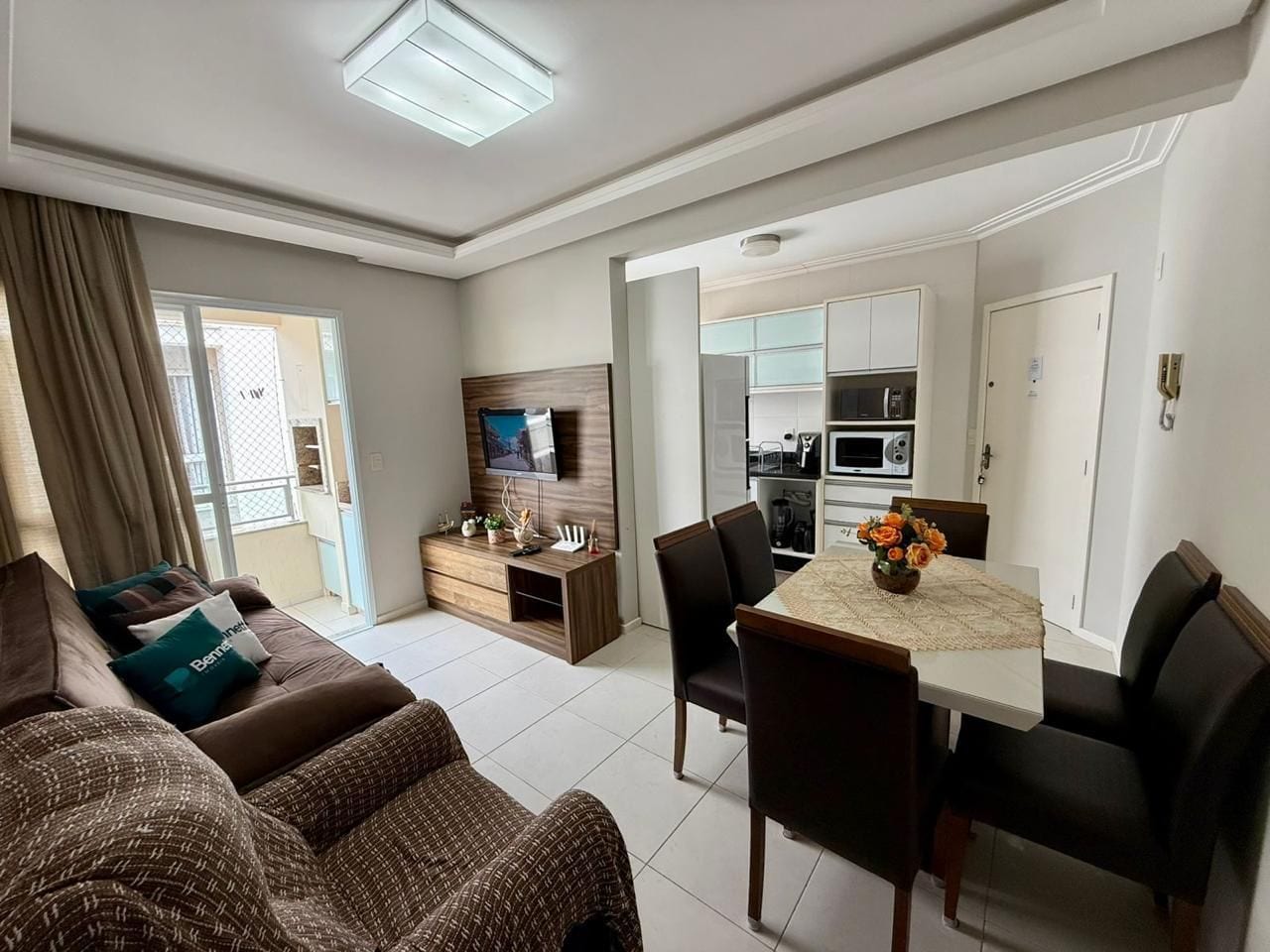 A modern living room with a sofa set, a dining table with chairs, a TV mounted on the wall, and a kitchen area visible through an open door.
