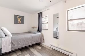 Ein Einzelbett mit grauer Bettdecke, ein an der Wand befestigtes Bild einer Hirschdackel, ein Fenster mit Blick und ein Spiegel.