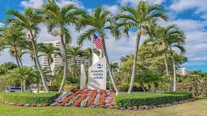 A South Seas Club sign with palm trees and a flag, surrounded by a landscaped garden. - Wake up to Clam Bay from this light- filled 2 bed 2 bath condo in South Seas Club on Marco Island (Marco Island)