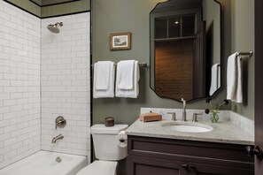 Une salle de bain avec des carreaux de céramique blancs de type « métro », une douche avec un pommeau de douche amovible, des toilettes, un lavabo avec un plan de travail en marbre et un miroir avec un cadre en bois.