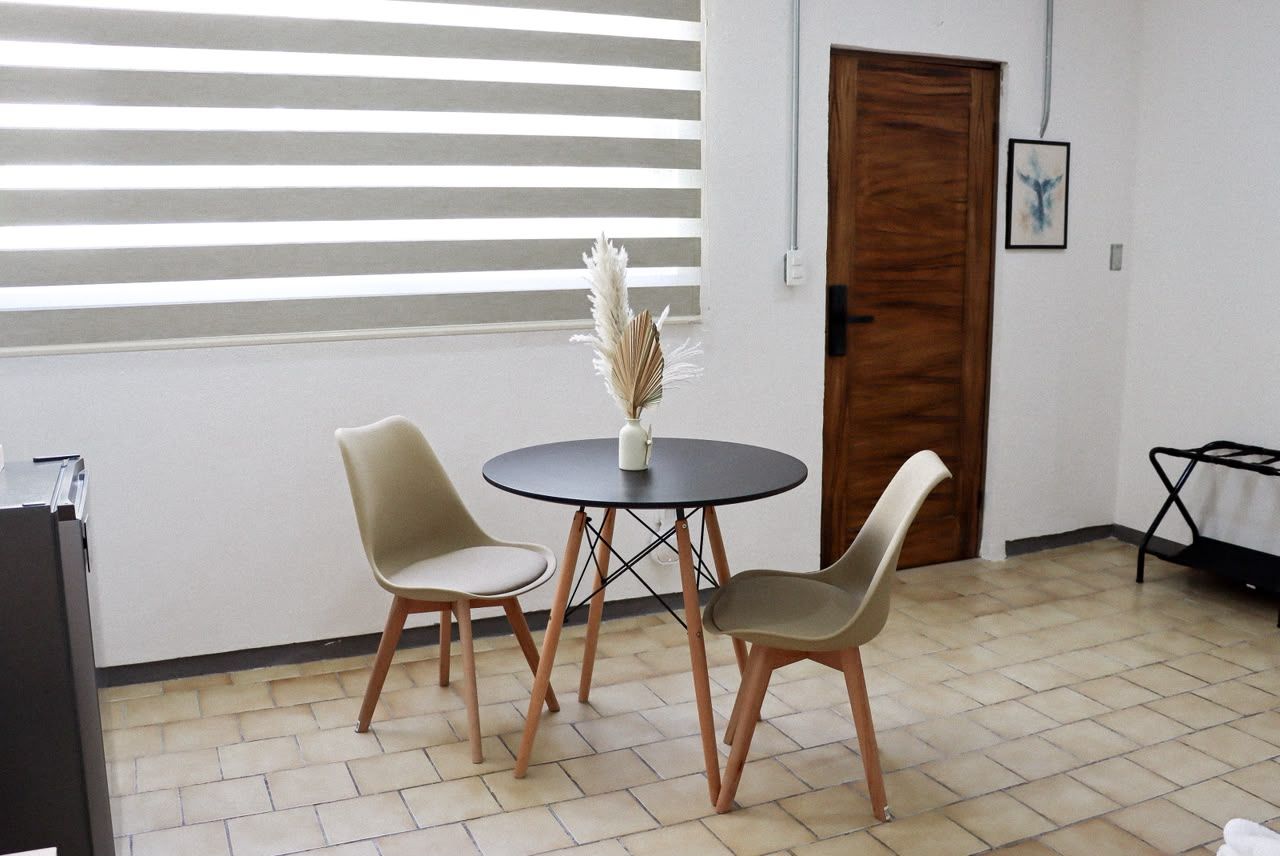 A room with a round table, two chairs, a vase with dried plants, a wooden door, and a window with blinds.