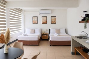 A hotel room with two beds, a small table, a chair, a desk with a sink, and artwork on the wall.