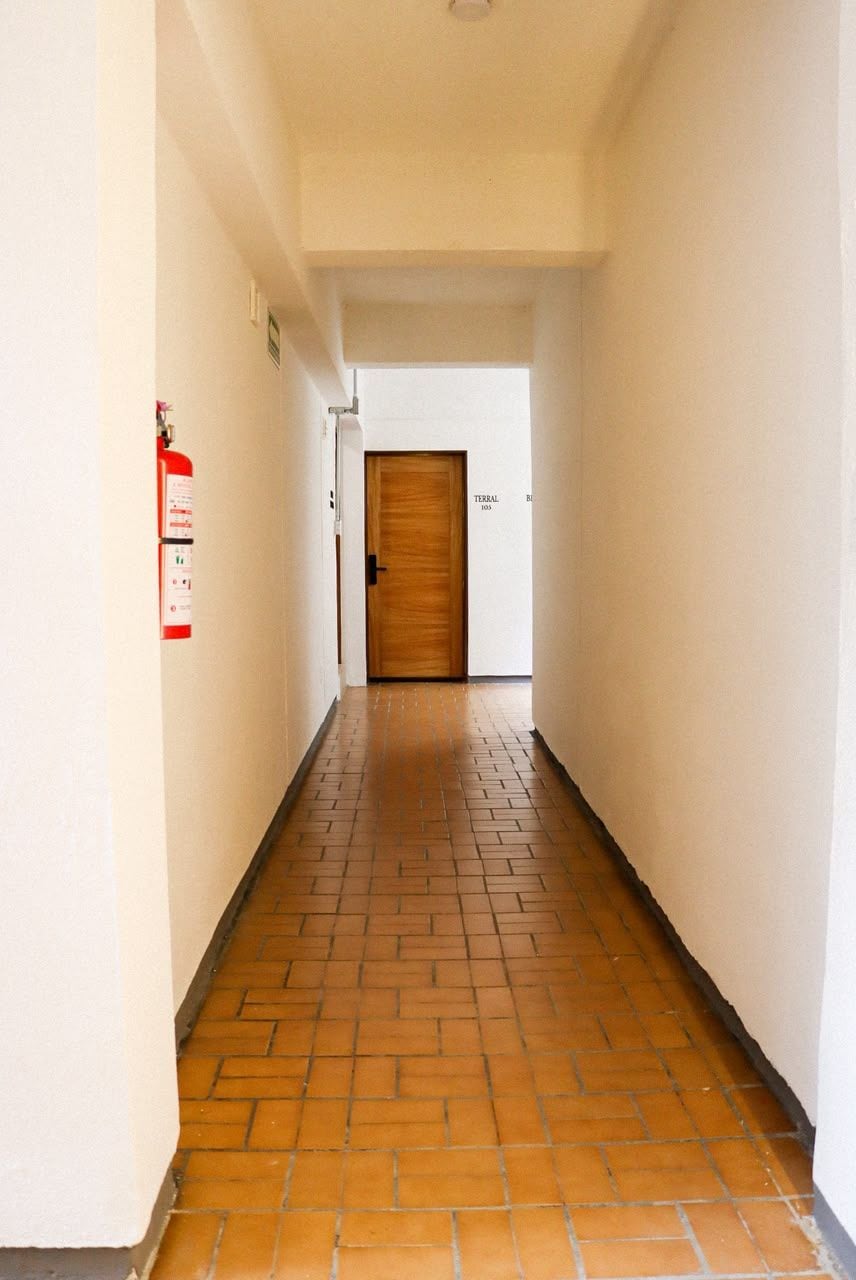 A corridor with a fire extinguisher on the wall, a wooden door at the end, and a tiled floor.