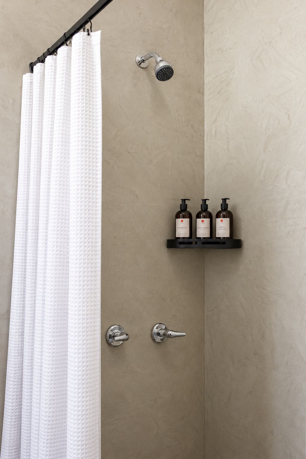 A bathroom with a shower curtain, a handheld showerhead, a shelf with three bottles, and two faucets.