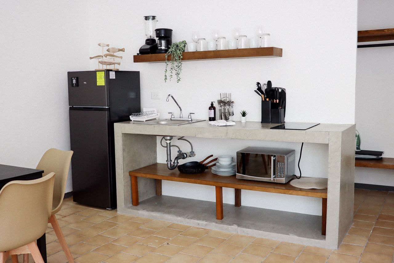 A compact kitchen with a built-in refrigerator, a microwave, and a sink. There is a wooden shelf above the counter holding kitchenware and a plant.