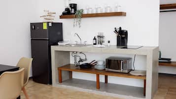 A compact kitchen with a built-in refrigerator, a microwave, and a sink. There is a wooden shelf above the counter holding kitchenware and a plant.