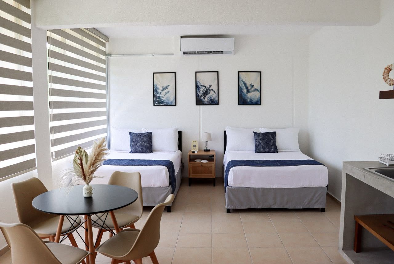 A hotel room with two beds, a dining table with chairs, a small kitchen area, and artwork on the wall.