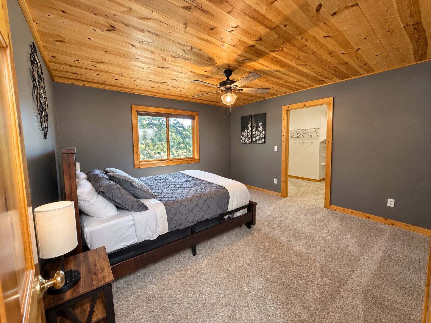 A bedroom with a wooden ceiling, a bed with a gray and white bedspread, a window, and a door leading to another room.