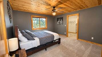 A bedroom with a wooden ceiling, a bed with a gray and white bedspread, a window, and a door leading to another room.