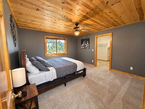 Un dormitorio con techo de madera, una cama con colcha gris y blanca, una ventana y una puerta que da a otra habitación.