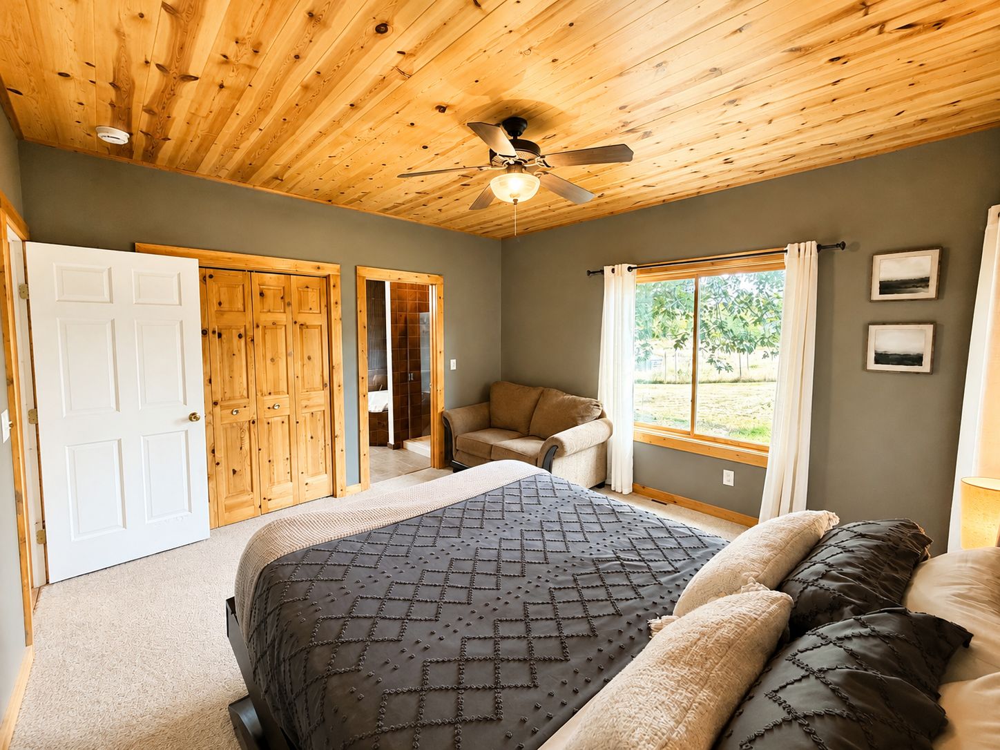 A bedroom with a wooden ceiling, a bed with patterned bedding, a window with curtains, and a door.