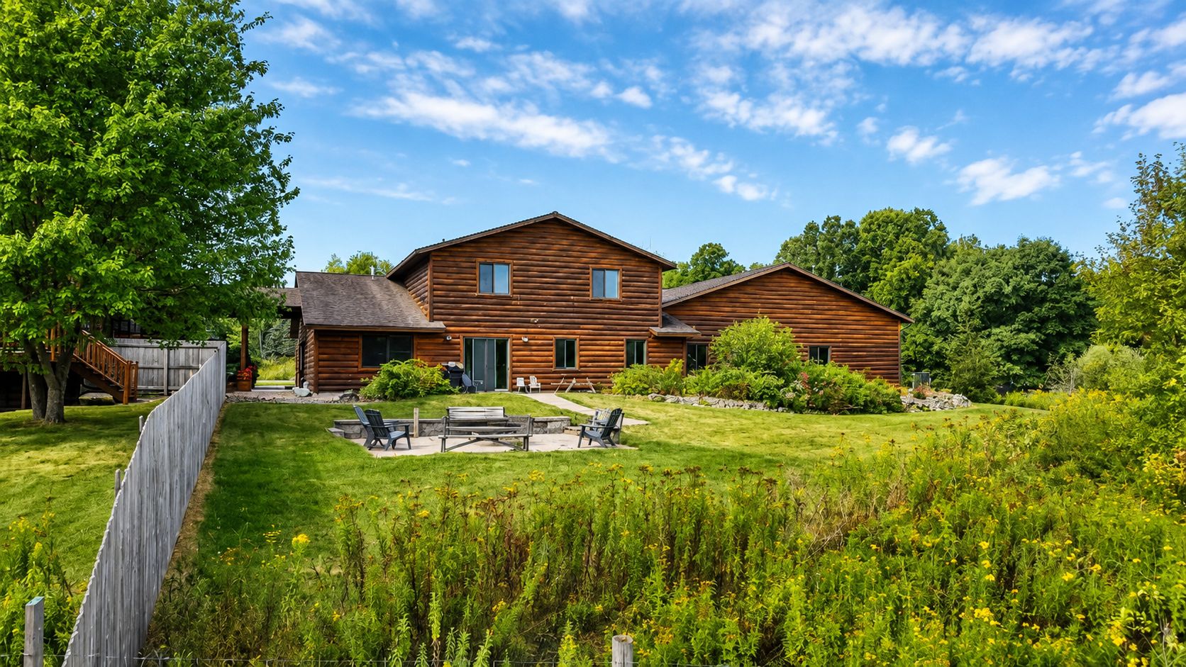 A wooden cabin with a patio area, surrounded by lush greenery and a clear sky.