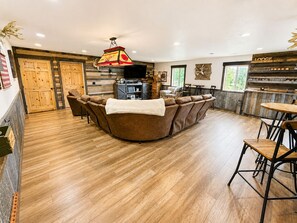 A cozy living area with a brown sofa, wooden flooring, a fireplace, and a bar area with a sign reading 'Lodge'.