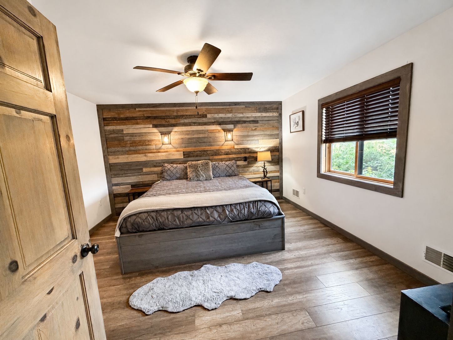A bedroom with a wooden headboard, a bed with a quilt, a ceiling fan, a window with blinds, and a rug on the floor.