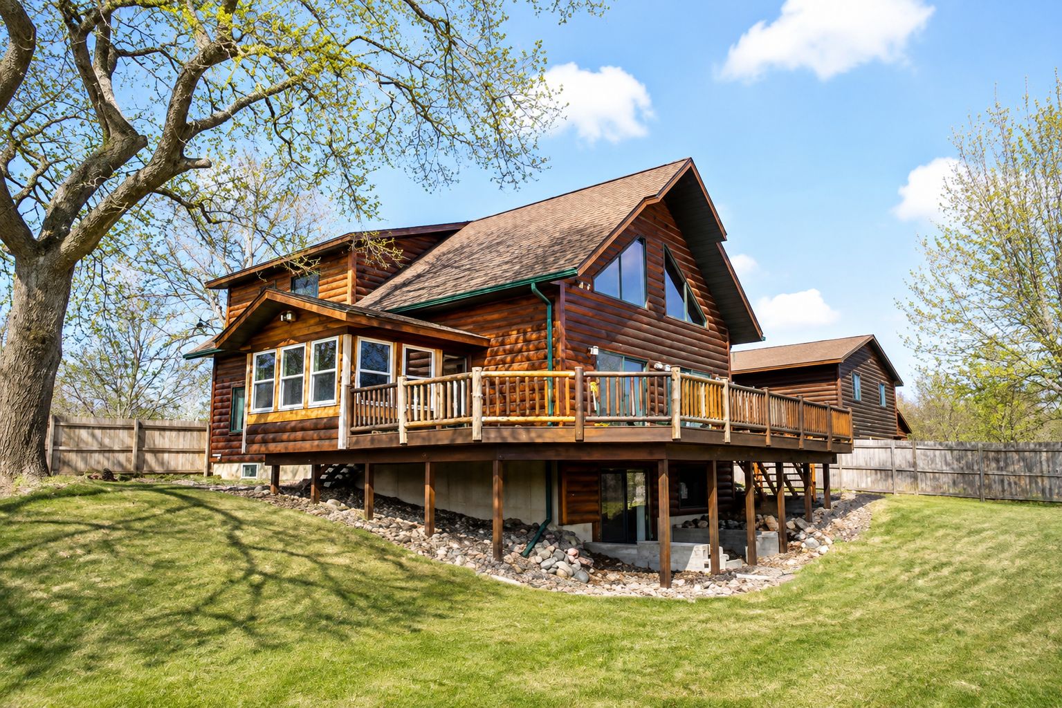 A two-story log cabin with a balcony, surrounded by a wooden deck and a stone foundation.