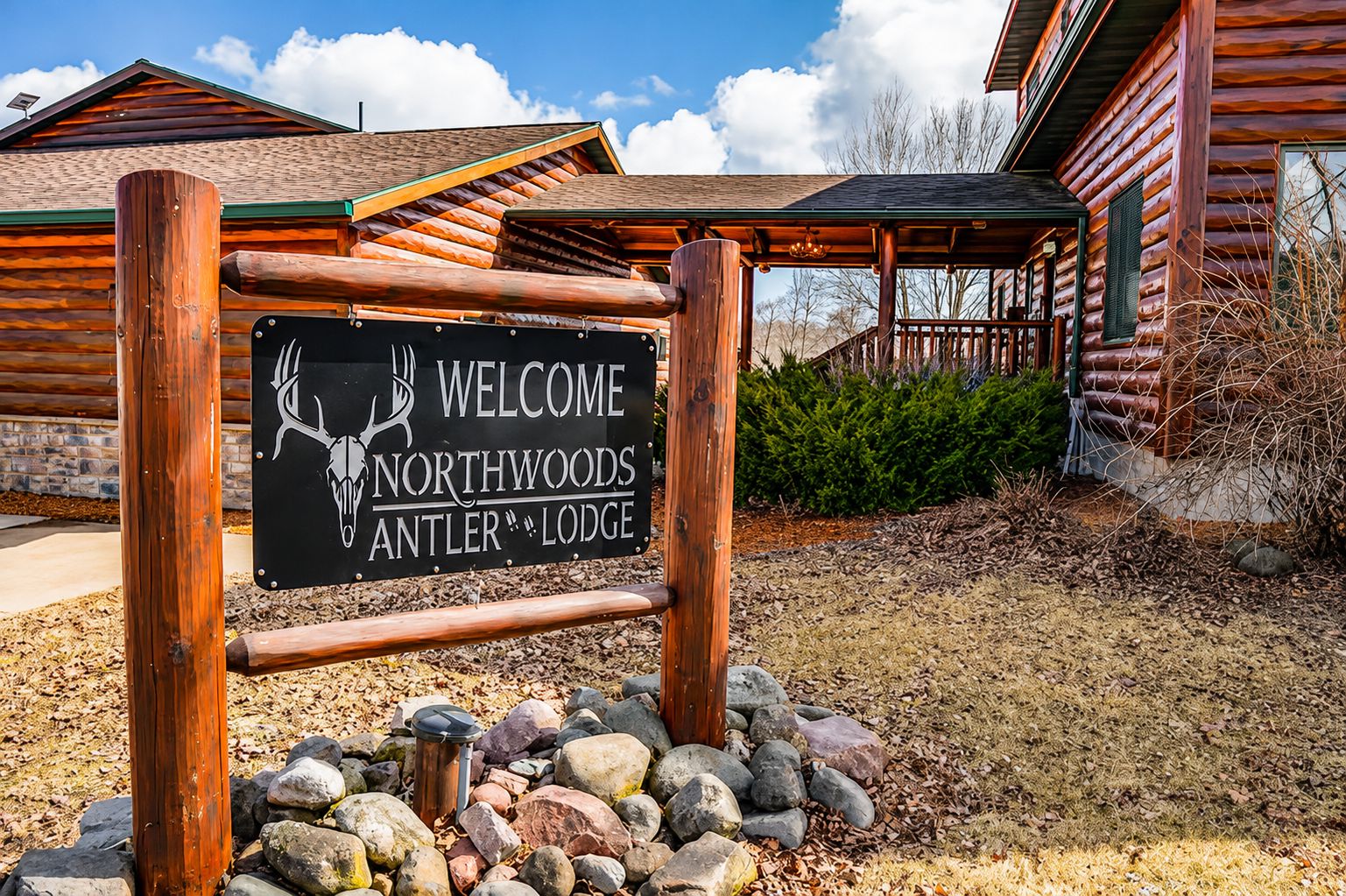 Cartell de benvinguda per a Northwoods Antler Lodge, amb una estructura de fusta rústica i fonaments de pedra.