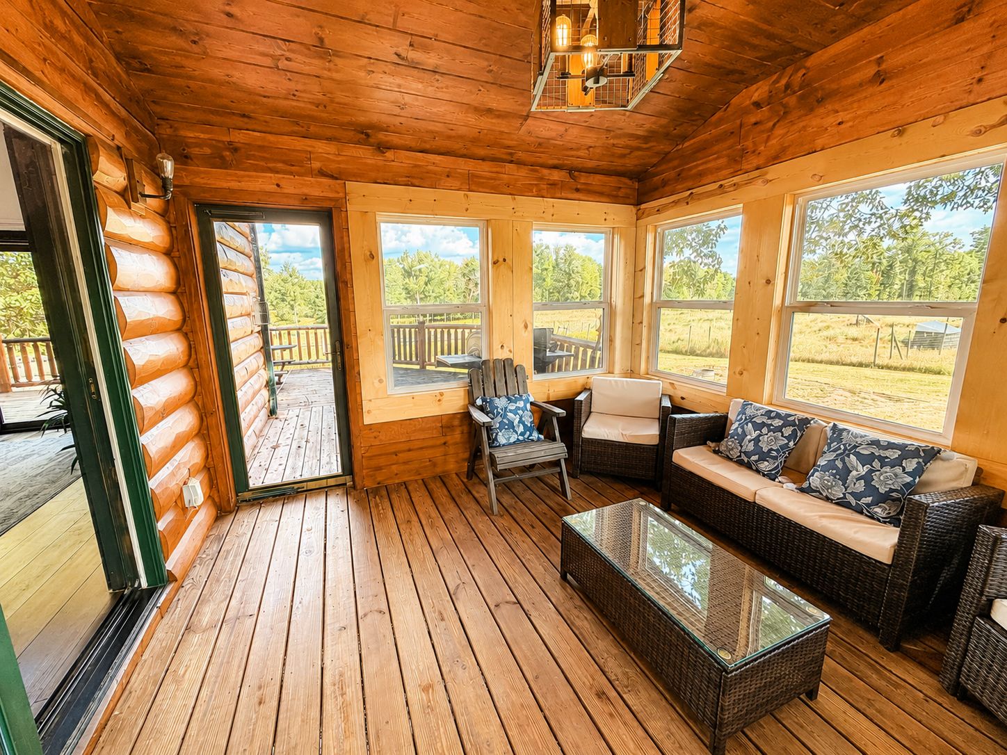 Interior de una cabaña de madera con piso de tablas, muebles de mimbre y ventanas amplias.