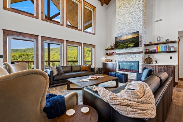 A spacious living room with a large window overlooking a green landscape, a fireplace, and a cozy seating area. - New! Loaded Luxury*Swim Spa*Theater*Games*Views! (Sevierville)