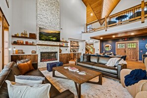 A spacious living area with a fireplace, leather sofas, a coffee table, and a high ceiling with wooden beams. - New! Loaded Luxury*Swim Spa*Theater*Games*Views! (Sevierville)