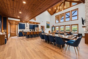 A spacious dining area with a long table and chairs, a kitchen with blue cabinets, and large windows offering views of the outdoors. - New! Loaded Luxury*Swim Spa*Theater*Games*Views! (Sevierville)